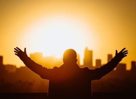 Silhouette of a man with his arms open wide as he is facing a city that's in the background with a giant light source in the middle of the sky