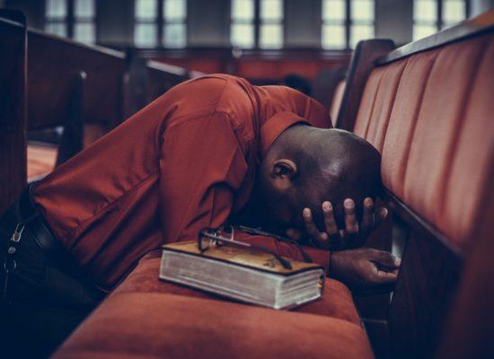 African American man on his knees praying with his head buried in his arms, located in between pew rows in a church that had glass windows in the background